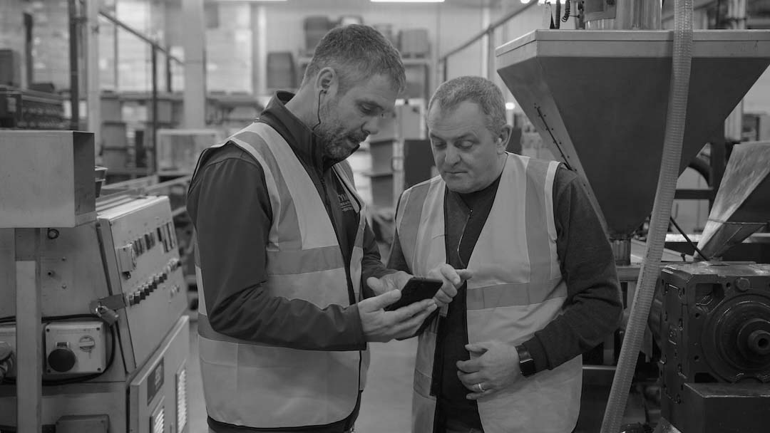 Two factory workers wearing high-visibility vests reviewing information on a smartphone beside industrial machinery.