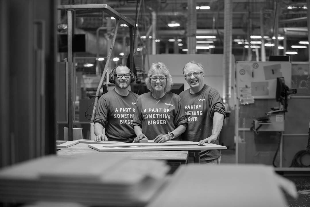 Three smiling factory employees standing together at a workbench in a manufacturing facility.