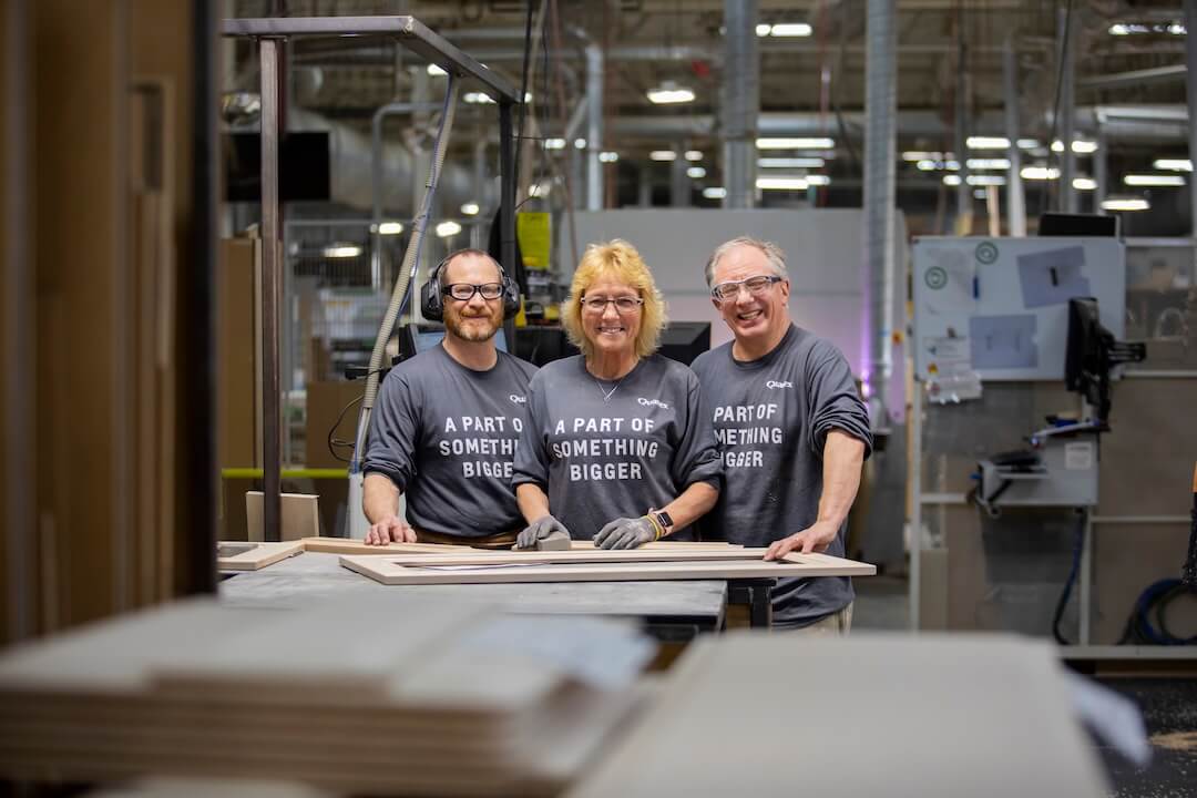 three smiling factory employees standing together at a workbench in a manufacturing facility.