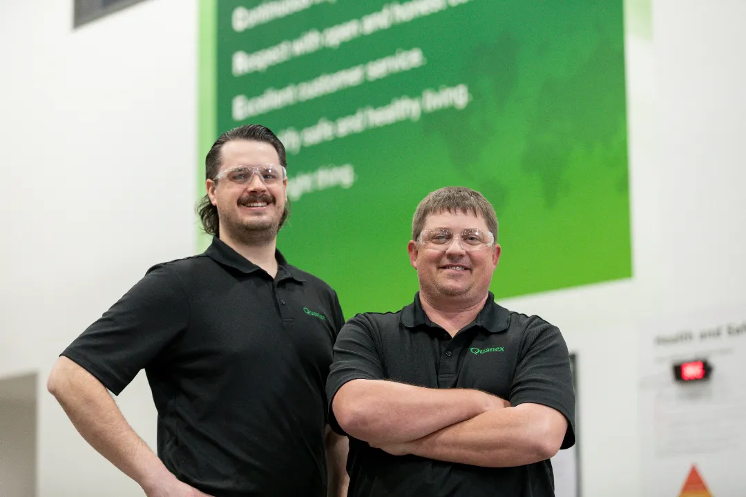 Two manufacturing employees wearing branded polo shirts and safety glasses inside a factory.