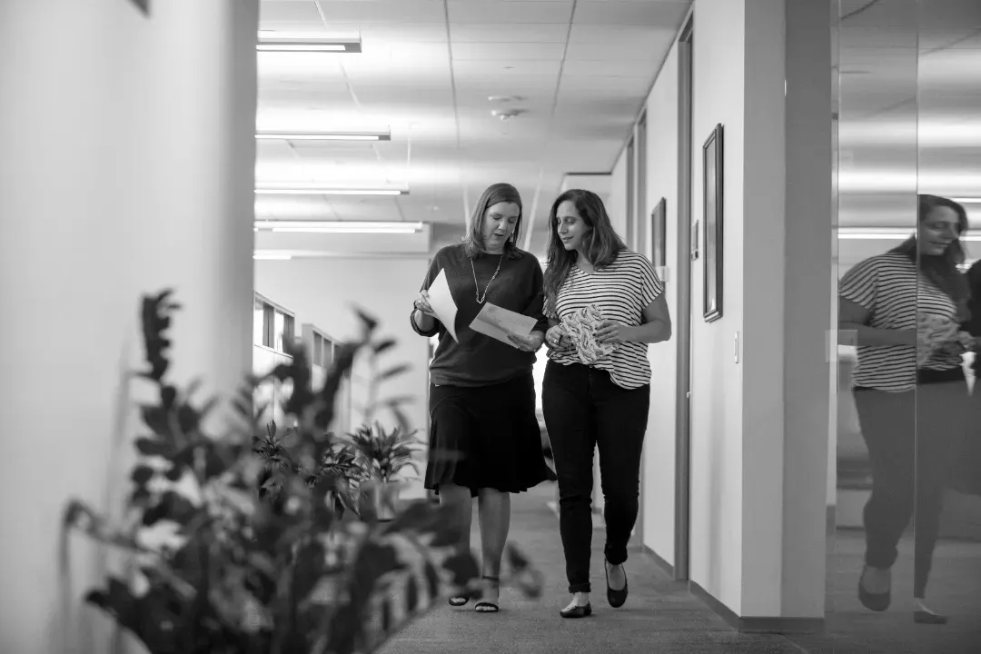 Two colleagues walking through an office corridor while reviewing documents together.