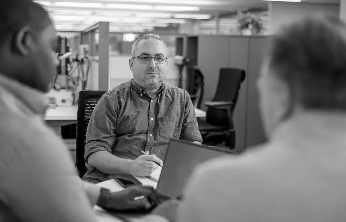 Three colleagues discussing work around a laptop in an open-plan office.