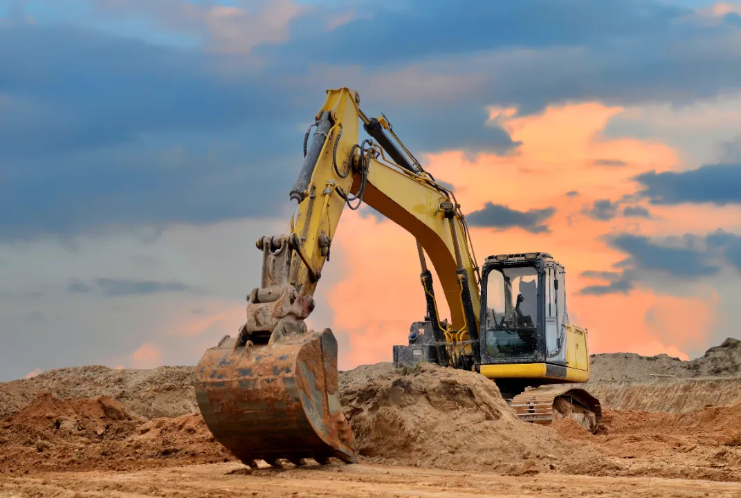Yellow tracked excavator digging on a construction site beneath a dramatic sunset sky.