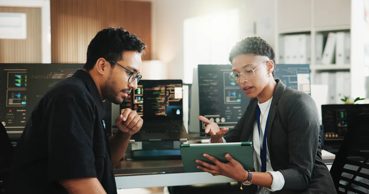 Two IT professionals reviewing information on a tablet beside computer screens displaying cybersecurity data and code.
