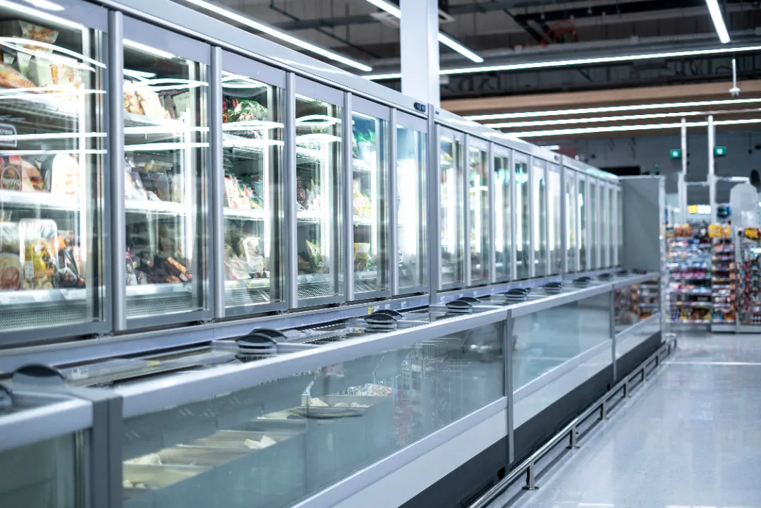 Long row of glass-door commercial freezers and open-top freezer units in a brightly lit supermarket aisle.